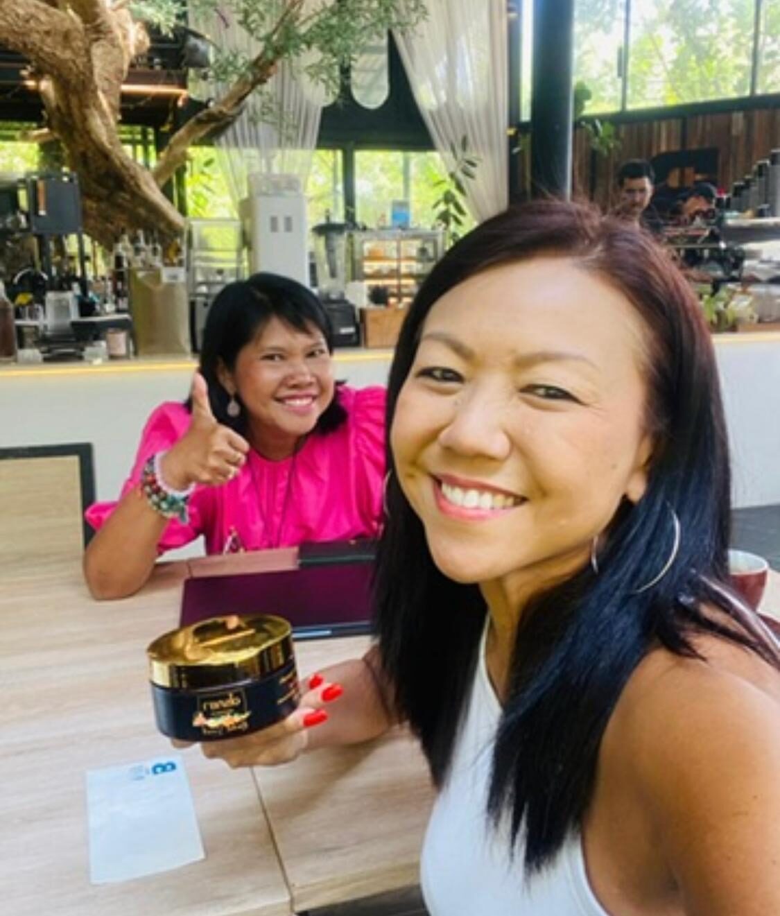 Two friends smiling together at a cafe in Thailand holding an Arokaya herbal scrub jar